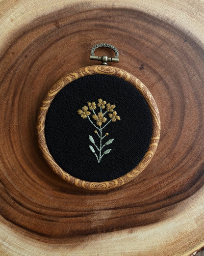 Embroidery hoop with floral design on black fabric, placed on a wooden surface.